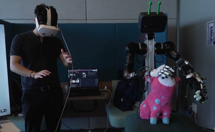 Dae wearing a Meta Quest headset, teleoperating an OpenArm bimanual robot at RLWRLD using only the headset's hand and arm tracking. The robot's hand rests on a plush toy; a laptop behind runs the teleoperation stack.